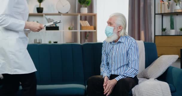 Attractive caring female doctor in protective mask visiting her senior male patient and attantivly listening about symptoms of his desease — Stock Video