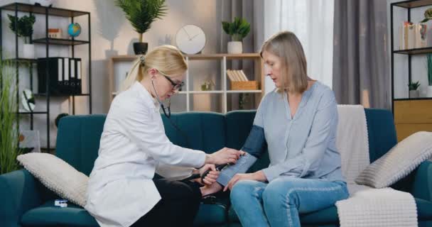 Pěkné sebevědomé kvalifikované blond žena-lékař měření krevního tlaku u nemocné ženy středního věku pomocí tonometru během plánované domácí návštěvy — Stock video
