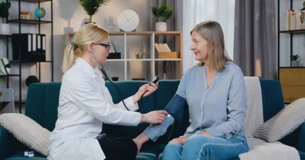 Trabajador médico calificado positivo agradable que se sienta en el sofá junto con paciente femenino y que mide la presión arterial usando el tonómetro durante visita casera — Vídeos de Stock