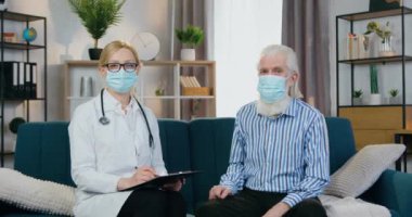 İyi görünümlü, kendine güvenen, deneyimli kadın doktor ve koruyucu maskeli kıdemli adam, doktor ev ziyareti sırasında kameranın önünde oturuyor.