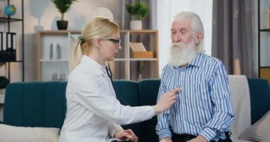 Yaşlı sakin sakallı adam kanepeye oturmuş, ev ziyareti sırasında kalp atışlarını dinleyen hoş bir bayan doktora bakıyor.