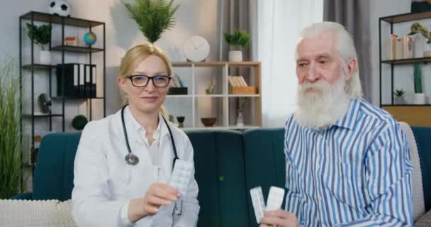 Vorderansicht der attraktiven, positiv ausgebildeten Ärztin und ihres männlichen Patienten, die auf der Couch in der Patientenwohnung sitzen und Tabletten in die Kamera präsentieren — Stockvideo
