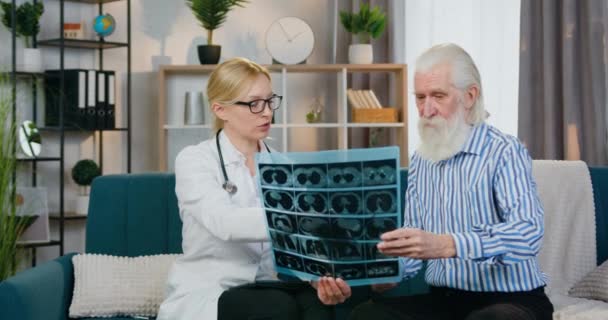 Attractive caring high-skilled female doctor revisioning results of x-ray image together with senior bearded male patient during visiting him at home — Stock Video