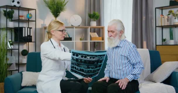 Całkiem uśmiechnięta doświadczona pracownica medyczna jest zadowolona ze skanu rentgenowskiego starszego mężczyzny i wita go z dobrymi wynikami podczas wizyty domowej — Wideo stockowe