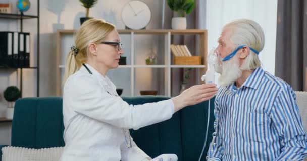 Agradable médico femenino de alta cualificación que se pone la máscara de oxígeno en la cara de los hombres para hacer la inhalación cuando lo visitan en casa, concepto de atención médica — Vídeo de stock