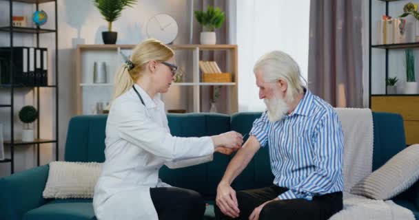 Vonzó pozitív gondoskodó szakképzett női egészségügyi dolgozó, aki otthoni látogatás során injekciót ad idős, ősz hajú szakállas férfinak — Stock videók
