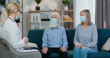 Keyifli ruh hali içinde, maskeli sarışın kadın doktor ev ziyareti sırasında koruyucu maskeler takan yaşlı çifti dinliyor.