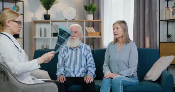 Εμφανίσιμη θετική ειδικευμένο φιλικό θηλυκό γιατρό εξηγώντας τα αποτελέσματα της ακτινογραφίας του γέρου γενειοφόρος ο οποίος κάθεται με τη σύζυγό του στον καναπέ κατά τη διάρκεια των γιατρών σπίτι επίσκεψη — Αρχείο Βίντεο
