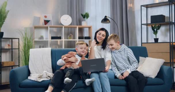 Bastante feliz 35 años de edad madre de pelo negro sentado en el sofá con sus tres sonrientes diferentes edades hijos durante el chat de vídeo con su pariente o marido — Vídeos de Stock