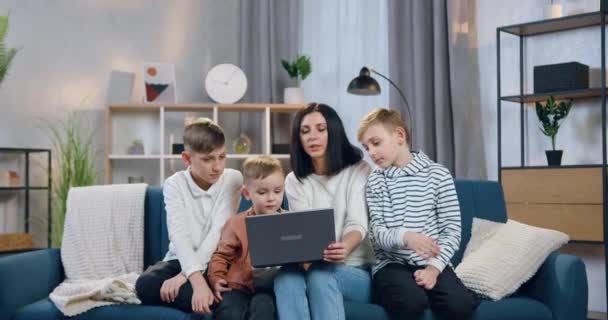 Atractiva sonrisa despreocupada joven madre explicando cómo utilizar el ordenador portátil de sus tres hijos de diferentes edades interesados cuando se sienta con ellos en el sofá suave en casa — Vídeo de stock