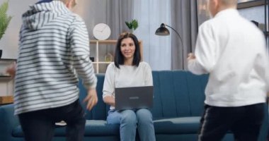 Güzel görünüşlü mutlu bir anne dizüstü bilgisayarla koltukta otururken üç farklı yaşta çocuk kucak kucak kucak ona koşuyor. Aile kavramı.