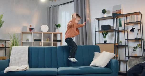 在当代公寓里，可爱快乐的小男孩一个人在柔软的沙发上跳着玩 — 图库视频影像