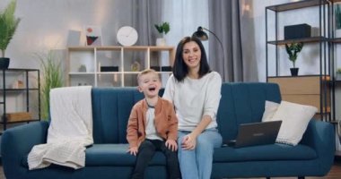 Sevimli, gülümseyen, siyah saçlı bir kadın, neşeli oğluyla birlikte evdeki kanepede oturuyor ve mutlu bir yüz ifadesiyle kameraya bakıyor.