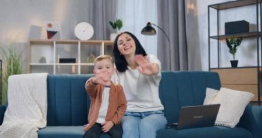 Sevimli gülümseyen mutlu annesi ve küçük neşeli oğlu evdeki kanepede oturup kameraya merhaba demek için el sallıyorlar.