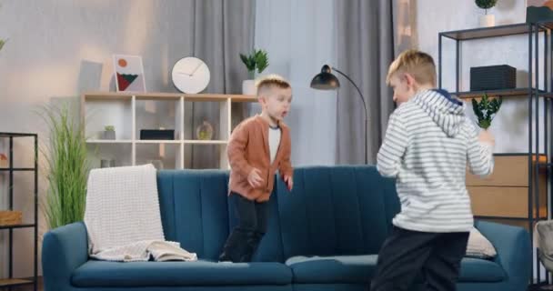 Härligt roliga 2-åriga och 8-åriga pojkar som har roligt tillsammans hemma, den minsta hoppar på soffan, den äldsta gör dansrörelser — Stockvideo