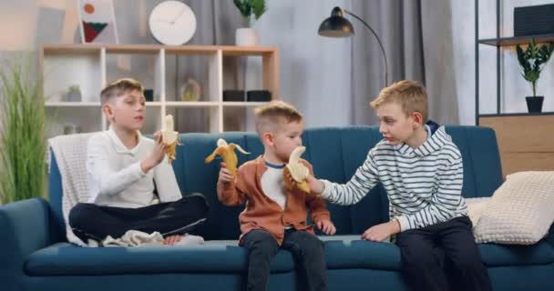 Schöne positive unterschiedlichen Alters drei Jungen sitzen auf der weichen Couch zu Hause und essen Bananen — Stockvideo