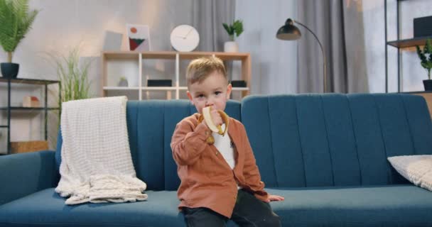행복 한 미소를 짓고 굶주린 작은 소년 이 집 소파에 앉아 잘 익은 바나나를 먹고 있는 모습 — 비디오