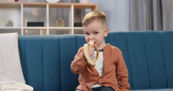 Netter kleiner zufriedener Junge schüttelt lustig den Kopf, wenn er reife Banane auf bequemem Sofa zu Hause isst — Stockvideo
