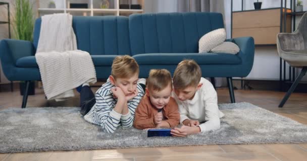 Lovely smiling happy different ages three boys lying on the floor and using smartphone to watch cartoon — Stock Video