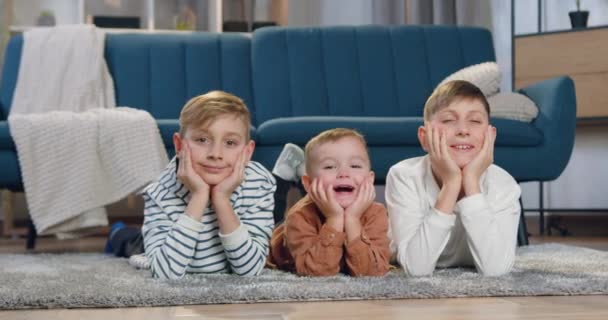 รูปภาพของเด็กชายสามคนที่มีความสุขที่ดูดี ยิ้มที่แตกต่างกันซึ่งนอนอยู่บนพื้นและมองไปที่กล้องเมื่อวางหัวของพวกเขาในมือ — วีดีโอสต็อก