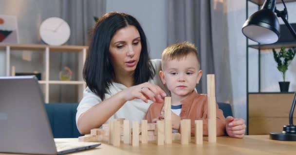 Vista frontal de una mujer de 30 años positiva y sin preocupaciones que ayuda a construir una torre de madera con bloques junto con su pequeño y lindo hijo — Vídeos de Stock