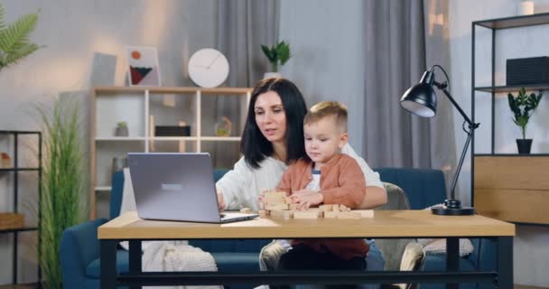 Goed uitziende glimlach gelukkig brunette werken op laptop op haar werkplek en vasthouden op knieën haar schattige kleine zoon die spelen met houten blokken — Stockvideo