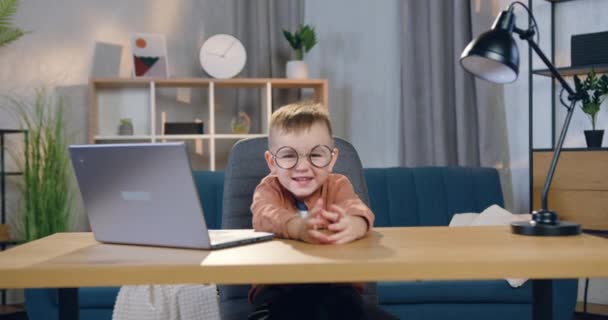 Прекрасний веселий смішний маленький хлопчик в окулярах сидить за столом з комп'ютером на ньому і показує великі пальці вгору, дивлячись на камеру — стокове відео