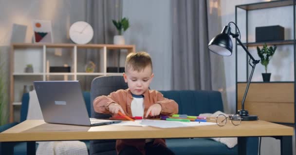 Портрет милого в хорошем настроении беззаботного маленького мальчика, который сидит за столом и рисует картинку фломастером — стоковое видео