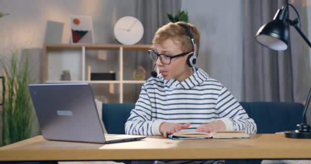 Atrakcyjny, skoncentrowany, inteligentny nastolatek w okularach i słuchawkach siedzący przed laptopem podczas lekcji online z nauczycielem i pytający o szczegóły zadania domowego — Wideo stockowe