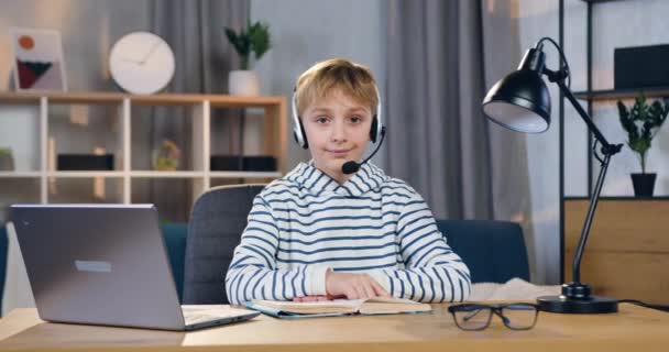 Close-up van knappe zelfverzekerde positieve slimme schooljongen in headset die zitten op zijn werkplek tijdens online les en kijken naar camera — Stockvideo