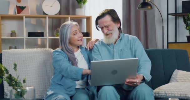 매력적 이고 행복 한 노부부는 집에 있는 소파에 앉아서 노트북 화면에서 온라인으로 읽은 웃긴 소식에 웃고 있습니다. — 비디오