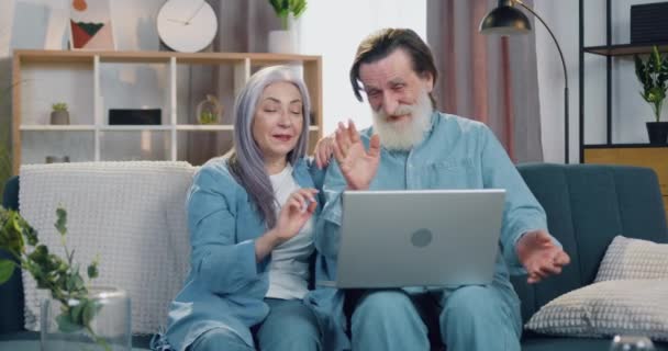 Adorable feliz sonriente pareja de ancianos sentados en el sofá en casa durante la videollamada con amigos usando la computadora y saludando con las manos para saludar y enviar besos al aire — Vídeos de Stock