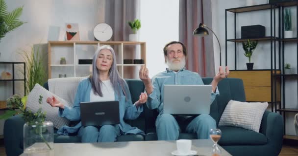 Attraktives entspanntes älteres Paar, das zu Hause auf einer weichen Couch mit Computern sitzt und mit geschlossenen Augen meditiert, um die Stille und Harmonie um sich herum zu genießen — Stockvideo