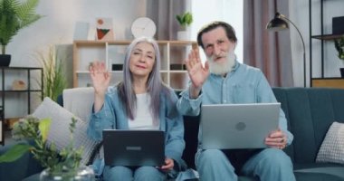 Çekici, gülümseyen, mutlu, yaşlı çiftin portresi. Evdeki kanepede oturmuş dizlerinin üstünde dizüstü bilgisayarlarını tutuyorlar ve kameraya el sallıyorlar.