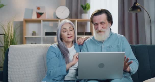 Goed uitziende positieve gepensioneerde echtpaar zitten op de bank in de hedendaagse woonkamer tijdens video gesprek met familieleden toepassen laptop — Stockvideo