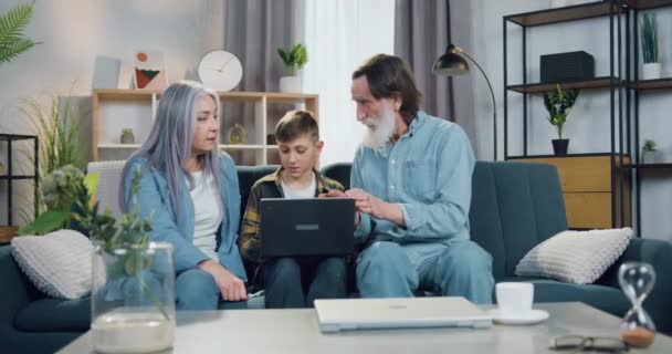 Привлекательный улыбающийся умный подросток, сидящий на диване между бабушкой и дедушкой и работающий над ноутбуком, пока они разговаривают друг с другом. — стоковое видео