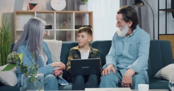 Pleasant kind caring mature grandmother and bearded grandfather helping with homework their lovely 10-aged grandson which sitting between them on the sofa and doing home task on laptop — Stock Video