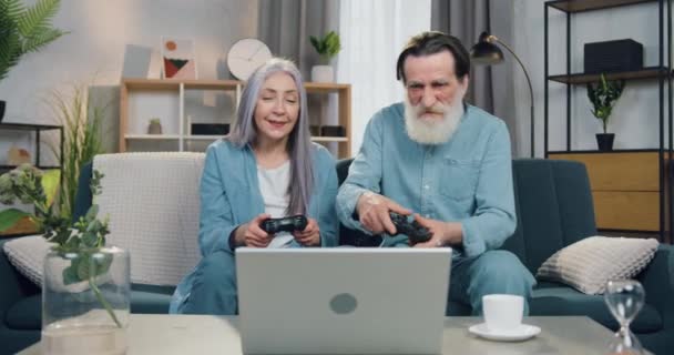 Concepto de ocio familiar donde la pareja de edad divertida y atractiva positiva se divierte en los juegos en el ordenador portátil utilizando joysticks, mujer celebrando la victoria cuando el hombre barbudo perdió el juego — Vídeos de Stock