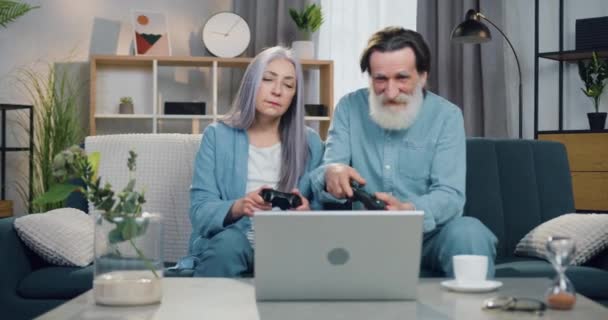 Vista frontal de los pensionistas masculinos y femeninos divertidos atractivos que se sientan juntos en el sofá en casa y juegan videojuegos en el ordenador portátil, el hombre ganó a su esposa y celebrar la victoria — Vídeo de stock
