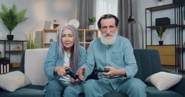 Portrét příjemného v dobré náladě veselý dospělý pár, který sedí na gauči doma a hraní pomocí joysticků, poté, co vyhrál dává vysoké pět navzájem — Stock video