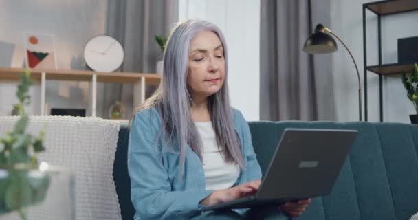 Aanzienlijke zelfverzekerde positieve gepensioneerde vrouw met lang grijs haar en casual kleding zitten op de bank thuis en werken aan laptop — Stockvideo