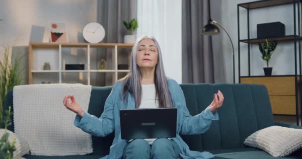 Atractiva mujer de cabello gris relajada sentada en el sofá y meditando con los ojos cerrados al hacer una pausa en su trabajo en el portátil — Vídeos de Stock