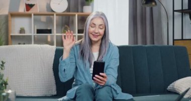 Çekici, mutlu, gülümseyen, gri saçlı 65 yaşında bir kadın evinde rahat bir koltukta oturuyor ve arkadaşıyla veya akrabasıyla cep telefonunda görüntülü sohbetin keyfini çıkarıyor.