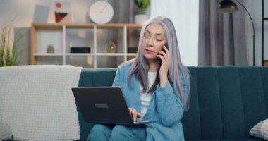 Kendine güveni yüksek 65 yaşında beyaz saçlı bir kadının portresi cep telefonuyla konuşan ve bilgisayarla modern oturma odası arka planında çalışan.