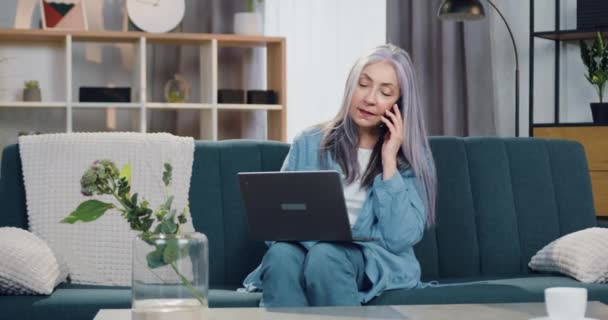 Mujer mayor de pelo gris feliz positiva atractiva que trabaja en el ordenador portátil en casa hablando simultáneamente en el teléfono inteligente, cámara lenta, vista frontal — Vídeos de Stock