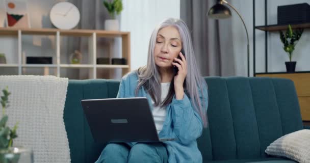 Kendine güveni tam, 60-65 yaşlarında bir kadın evdeki kanepede oturuyor ve cep telefonu konuşmaları sırasında bilgisayarda çalışıyor. — Stok video
