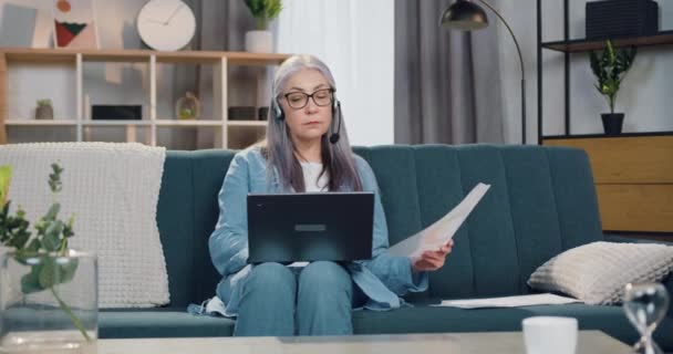 El trabajo remoto desde el concepto de casa donde atractiva mujer de 65 años de edad, de pelo gris y confianza seria en los auriculares sentados en el sofá y trabajando en el ordenador portátil — Vídeo de stock