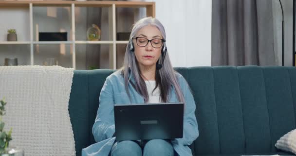 Atractiva mujer mayor de pelo gris con éxito y confianza en los auriculares sentada frente a la computadora portátil durante la videoconferencia y discutiendo el informe con gráficos con compañeros de trabajo — Vídeos de Stock