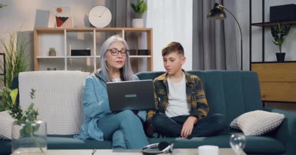Красивый улыбающийся удовлетворенный 10-летний мальчик сидит на диване вместе со своей милой добросердечной седой бабушкой и они используют ноутбук — стоковое видео