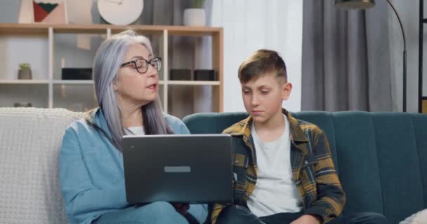 Precioso tipo experimentado abuelita de edad avanzada en gafas sentado en el sofá junto con su apuesto nieto positivo y explicándole la tarea en el ordenador — Vídeos de Stock